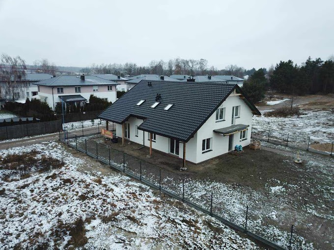 Nowoczesny, parterowy dom z ciemną dachówką, białymi ścianami i elementami drewnianymi, otoczony metalowym ogrodzeniem na działce pokrytej śniegiem.