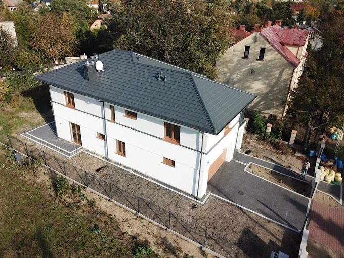 Dwukondygnacyjny dom murowany z szarą dachówką, brązowymi oknami i garażem wbudowanym, widok z lotu ptaka, z wybrukowanym podjazdem i częściowo ogrodzonym terenem.