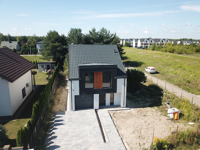 Nowoczesny dom jednorodzinny z płaskim dachem i garażem, wykończony w bieli i czerni, z brukowanym podjazdem i niezagospodarowanym terenem wokół, widziany z góry.