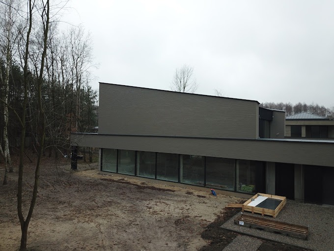 Nowoczesny budynek z płaskim dachem i dużymi oknami, widok z góry na plac budowy z drewnianymi paletami, szary odcień elewacji i ponure niebo.