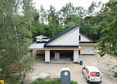 Surowy stan budynku z białego bloczka z charakterystycznym skośnym dachem i otwartą przestrzenią garażową, widok z góry, otoczony zielenią drzew.