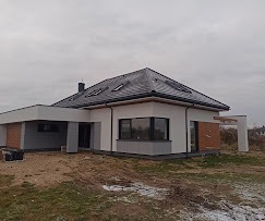 Parterowy dom z szarą dachówką i elementami dekoracyjnymi z drewna na elewacji, widoczny w pochmurny dzień.