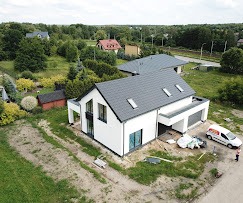 Nowoczesny dom murowany z dwuspadowym dachem z szarej dachówki, widok z drona, zarysowana działka, biała elewacja, częściowo odsłonięte okna, zaparkowany biały bus z logo firmy w tle.