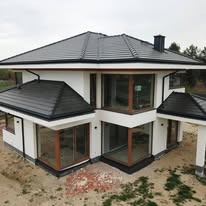 Dwukondygnacyjny dom z ciemną dachówką, jasną elewacją i dużymi oknami narożnymi. Na pierwszym planie gruz budowlany. Widok z góry.