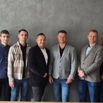 Zespół budowlany: pięciu mężczyzn w swobodnych strojach stoi na tle szarej ściany, uśmiechając się do kamery. Widoczne detale ubioru i fryzur.