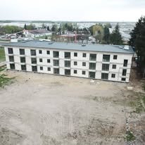 Dwukondygnacyjny, jasny budynek w stanie surowym zamkniętym, z widocznymi otworami okiennymi i drzwiami balkonowymi, na tle niezagospodarowanego terenu.