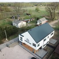 Nowoczesny, dwukondygnacyjny dom z białymi ścianami i grafitowym dachem, widziany z lotu ptaka na tle zielonego terenu. Minimalistyczna architektura, płaski dach.