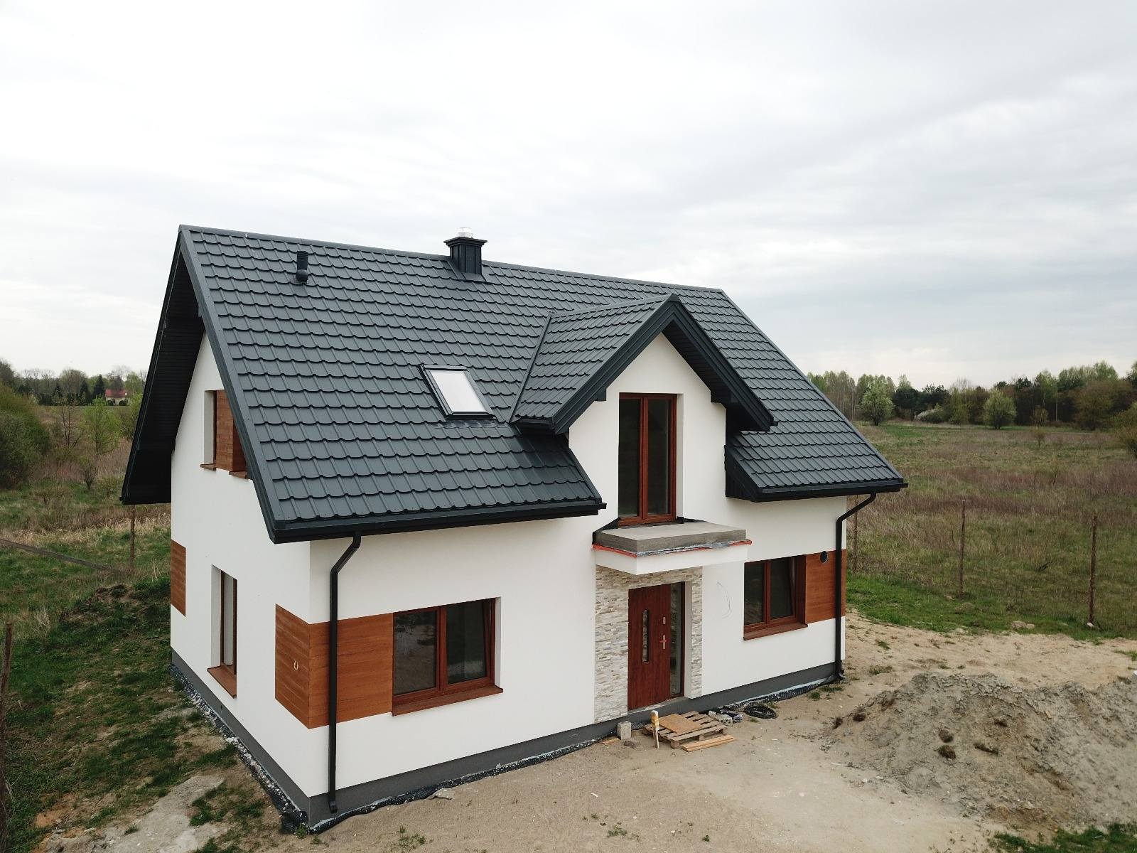 Nowo wybudowany, dwukondygnacyjny dom z ciemnoszarą dachówką, białymi ścianami i drewnianymi akcentami, otoczony ziemią i zielenią, w pochmurny dzień.