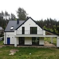 Nowoczesny dom z białymi ścianami i czarnym dachem, balkon z metalową barierką, niebieskie drzwi wejściowe, otoczony zielenią, las w tle.