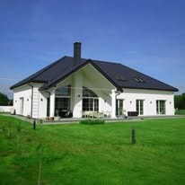 Elegancki, parterowy dom z białymi ścianami i ciemnym dachem, otoczony zielonym trawnikiem. Widoczne duże okna i komin. Jasne, słoneczne ujęcie.