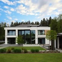 Nowoczesny, dwukondygnacyjny dom z płaskim dachem, dużymi przeszkleniami i jasną elewacją na tle zieleni i błękitnego nieba z chmurami. Minimalistyczny ogród.