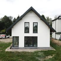 Nowoczesny, biały dom jednorodzinny z ciemnymi oknami i dachem dwuspadowym na tle drzew. Minimalistyczny design, świeżo wykończona elewacja, widoczny fragment samochodu dostawczego.