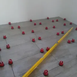 Ułożone szare płytki podłogowe z czerwonymi klipsami poziomującymi i żółtą poziomicą w rogu pomieszczenia.