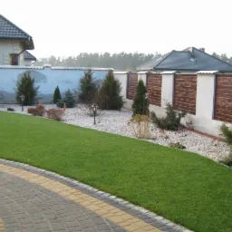 Ogród z nową kostką brukową na podjeździe, równo przystrzyżonym trawnikiem i ozdobnym ogrodzeniem z drewnianymi panelami w tle.