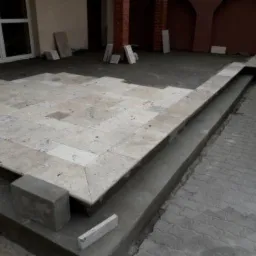 Układanie jasnych, kwadratowych płyt kamiennych na tarasie z widocznymi etapami prac i niedokończonymi fragmentami, częściowo przykrytymi szarym cementem. Widoczne ceglane filary i okno w tle.