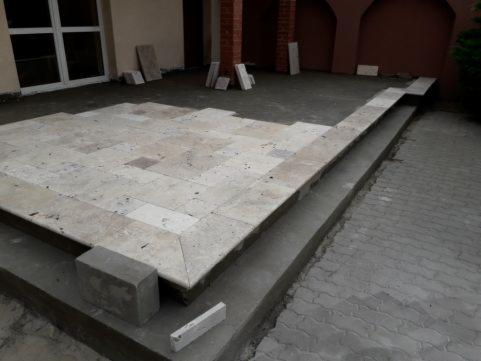 Układanie jasnych, kwadratowych płyt kamiennych na tarasie z widocznymi etapami prac i niedokończonymi fragmentami, częściowo przykrytymi szarym cementem. Widoczne ceglane filary i okno w tle.