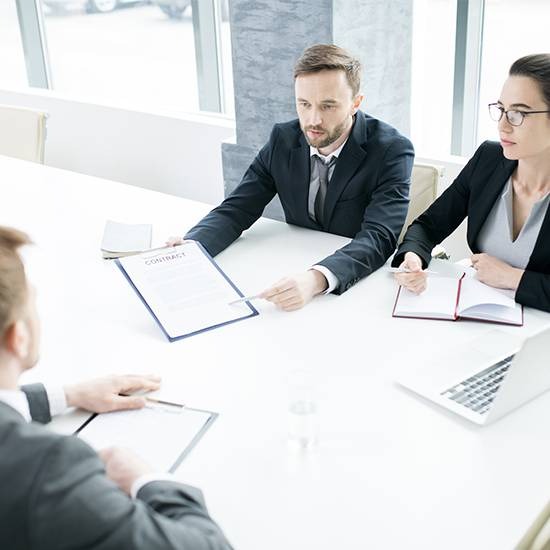 Spotkanie biznesowe przy białym stole: mężczyzna w garniturze prezentuje umowę, kobieta w okularach notuje, widoczny laptop i szklanka wody.