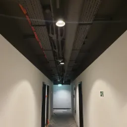 Korytarz biurowy w trakcie prac remontowych, podłoga zabezpieczona folią, widoczne kable w korytkach pod sufitem i świeżo pomalowane ściany.