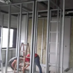 Montaż metalowego stelaża ścian działowych z widoczną izolacją z wełny mineralnej, dwóch pracowników przy pracy, drabina drewniana i aluminiowa w pomieszczeniu z oknami.
