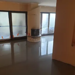 Świeżo wylana, błyszcząca posadzka w pomieszczeniu w trakcie remontu w Pile, okna zabezpieczone folią, widoczny kominek.