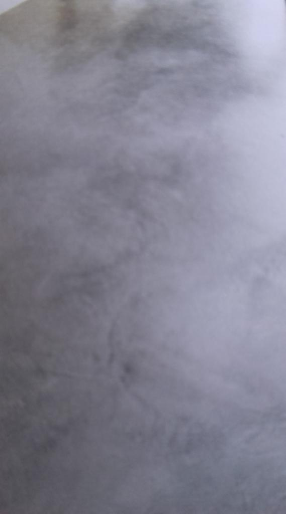 Szary, nierównomierny wzór na powierzchni, przypominający strukturę betonu lub tynku dekoracyjnego z delikatnymi smugami i cieniowaniem.