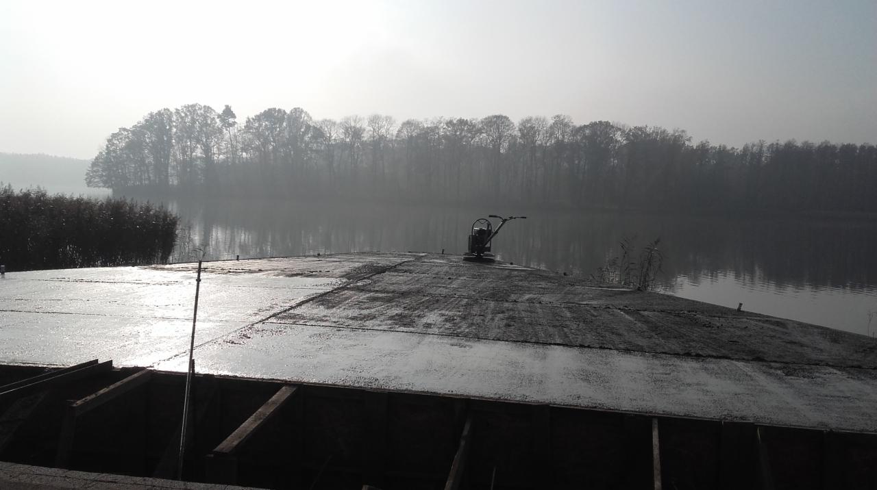 Świeżo wylany jastrych na tle jeziora w porannej mgle, widoczna maszyna do zacierania.