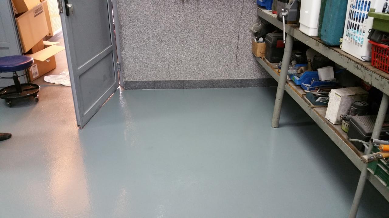 Jasnoszara, gładka posadzka żywiczna w pomieszczeniu gospodarczym z regałem narzędziowym i otwartymi drzwiami. Ściany wykończone w szarym, nakrapianym tynku.