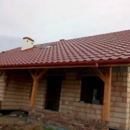 Budowa domu z czerwonym dachem w Budzisławiu Kościelnym, widoczne drewniane elementy konstrukcyjne i ceglane ściany w trakcie wznoszenia.