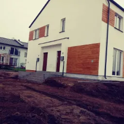 Nowoczesny, biały bliźniak z elementami dekoracyjnymi z paneli drewnopodobnych na elewacji, widoczne schody wejściowe i przygotowany teren wokół budynku.