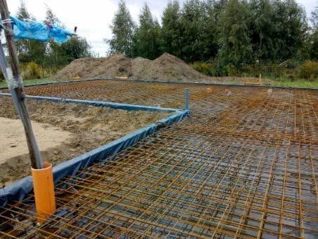 Przygotowanie zbrojenia fundamentów pod dom jednorodzinny, widoczne siatki zbrojeniowe, szalunki z folii oraz rury instalacyjne, w tle hałda ziemi.