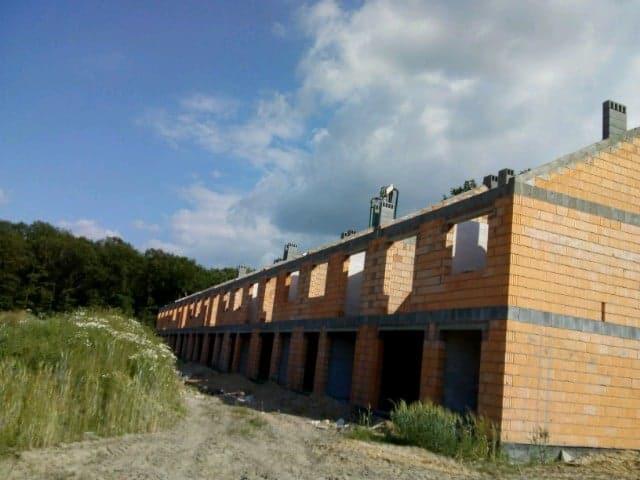 Surowy stan szeregu domów w trakcie budowy z czerwonej cegły, widoczne otwory okienne i drzwiowe, teren wokół niezagospodarowany, pochmurne niebo.