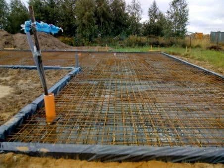 Przygotowanie zbrojenia pod fundament płyty domu murowanego z widocznym szalunkiem i wystającymi rurami instalacyjnymi.