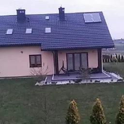 Gotowy dom jednorodzinny z kremową elewacją, ciemną dachówką i panelami słonecznymi na dachu, otoczony trawnikiem i tujami.