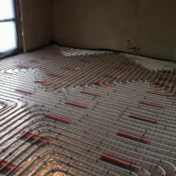 Białe rury ogrzewania podłogowego ułożone spiralnie na czerwonej siatce w jasnym pomieszczeniu, widok z g&oacute;ry.