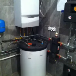 Biały piec gazowy Vaillant z podłączonymi rurami w pomieszczeniu wyłożonym szarymi płytkami. Widoczne elementy instalacji grzewczej i hydraulicznej.