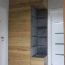 Drewniana zabudowa z wnęką wyłożoną szarą tapicerką, tworzącą siedzisko w przedpokoju, obok białe drzwi