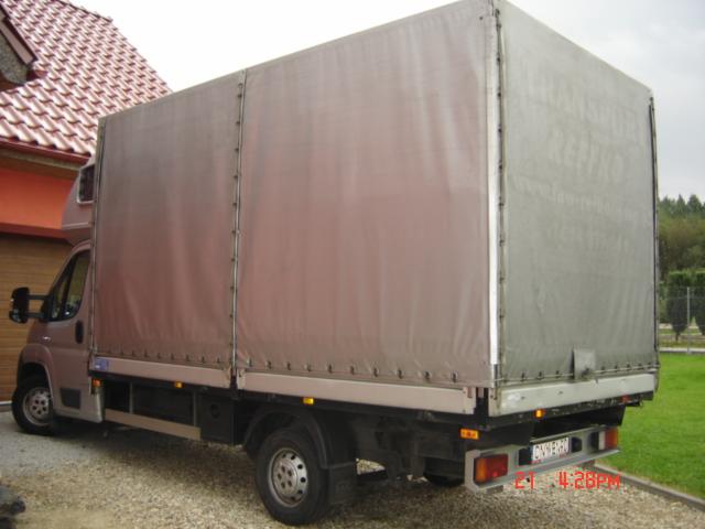 Widok z tyłu szarego busa z plandeką, zaparkowanego na żwirowym podjeździe przed domem z czerwoną dachówką i zielonym trawnikiem w tle.