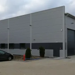 Nowoczesna hala przemysłowa z szarej płyty warstwowej, z czarnymi akcentami u dołu, widoczna brama wjazdowa i okna, przed budynkiem zaparkowane samochody osobowe i hydrant.