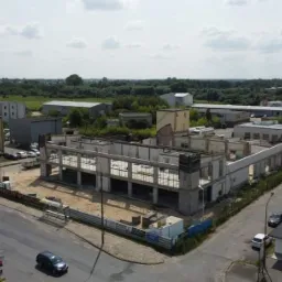 Budowa hali z żelbetu w trakcie realizacji, widok z drona na plac budowy z widocznymi elementami konstrukcyjnymi i zbrojeniami, w tle zabudowa miejska.