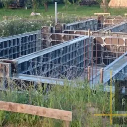 Szare szalunki fundamentowe z widoczną konstrukcją na trawiastym terenie, przygotowane do zalania betonem, w tle zieleń i zabudowania gospodarcze.