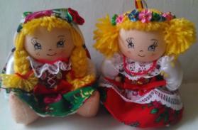 Dwie ręcznie robione lalki w strojach ludowych, z blond włosami i kwiatowymi wiankami, siedzą obok siebie na jasnym tle. Detaliczne wzory na ubraniach.