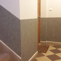 Klatka schodowa z wykończonymi ścianami w szarym, nakrapianym odcieniu, kontrastująca z białymi górnymi partiami ścian i brązowo-beżową szachownicą na podłodze, widoczne schody z beżowymi płytkami...