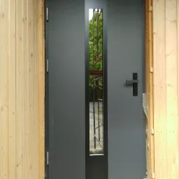 Grafitowe drzwi zewnętrzne z wąskim, pionowym przeszkleniem, osadzone w surowej ramie drewnianej, widoczne rusztowanie za szybą.