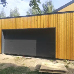 Nowoczesna, segmentowa brama garażowa w kolorze grafitowym, z drewnianą elewacją w pionowe deski, widoczna paleta drewniana na ziemi