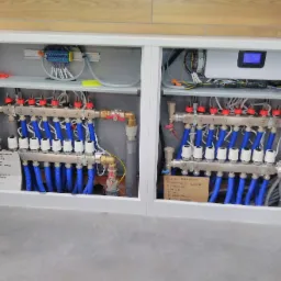 Rozdzielacz ogrzewania podłogowego z niebieskimi rurami w szafce, z numeracją od 1 do 8, widoczne także elementy elektryczne i notatka odręczna z rozpiską pomieszczeń.