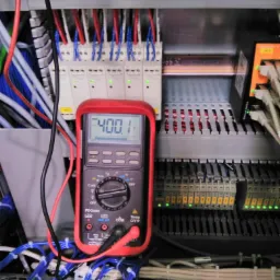 Multimetr z czerwonym obramowaniem pokazuje wartość 400.1 na tle szafy sterowniczej z licznymi przewodami i złączkami.