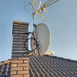 Antena satelitarna i antena telewizyjna zamontowane na kominie z cegły klinkierowej na dachu pokrytym dachówką ceramiczną, widok z dołu na tle błękitnego nieba.
