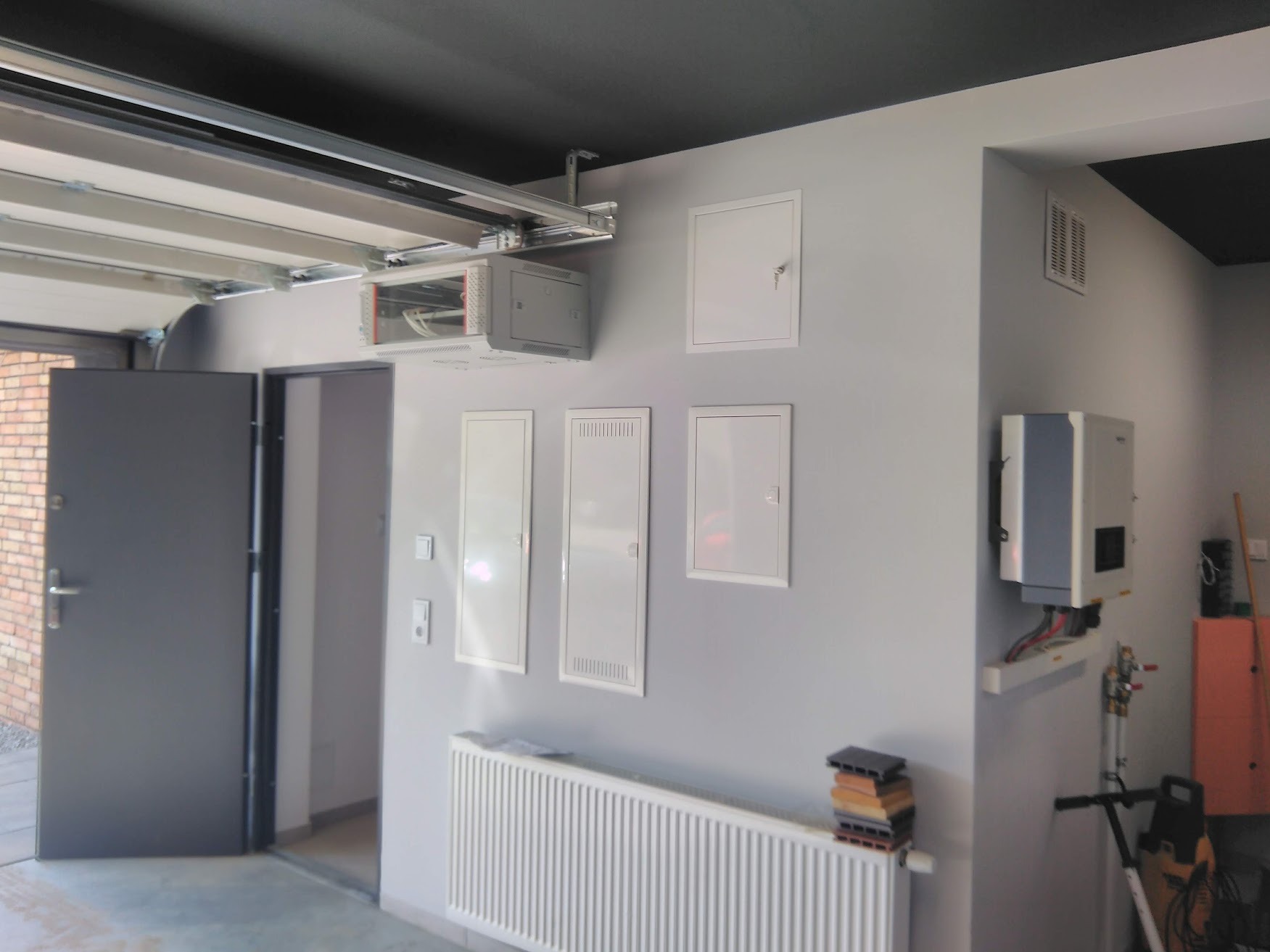 Wnętrze garażu z zamontowaną szyną sufitową dla bramy, trzema białymi szafkami elektrycznymi na ścianie, białym grzejnikiem i falownikiem solarnym.
