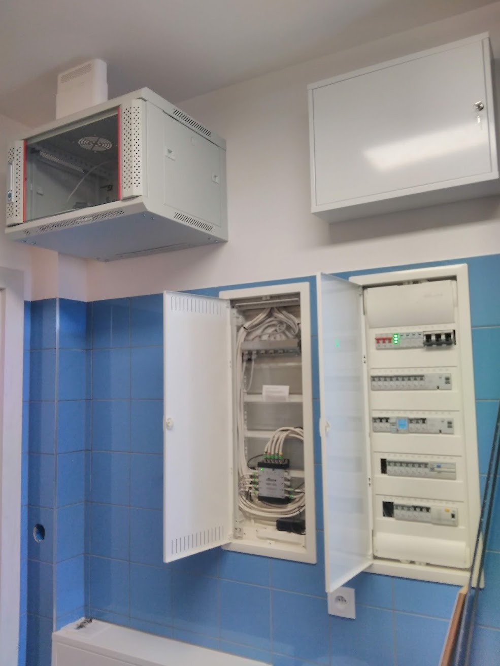 Rozdzielnica elektryczna z otwartymi drzwiczkami, szafa rack pod sufitem i dodatkowa skrzynka na ścianie z niebieską glazurą.