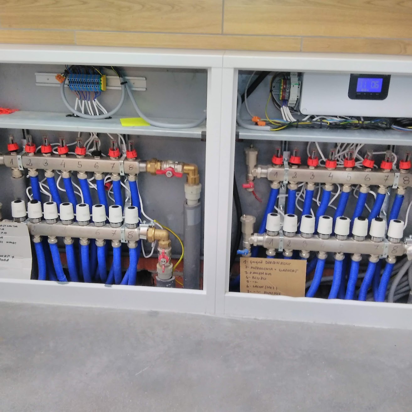 Rozdzielacz ogrzewania podłogowego z niebieskimi rurami w szafce, z numeracją od 1 do 8, widoczne także elementy elektryczne i notatka odręczna z rozpiską pomieszczeń.